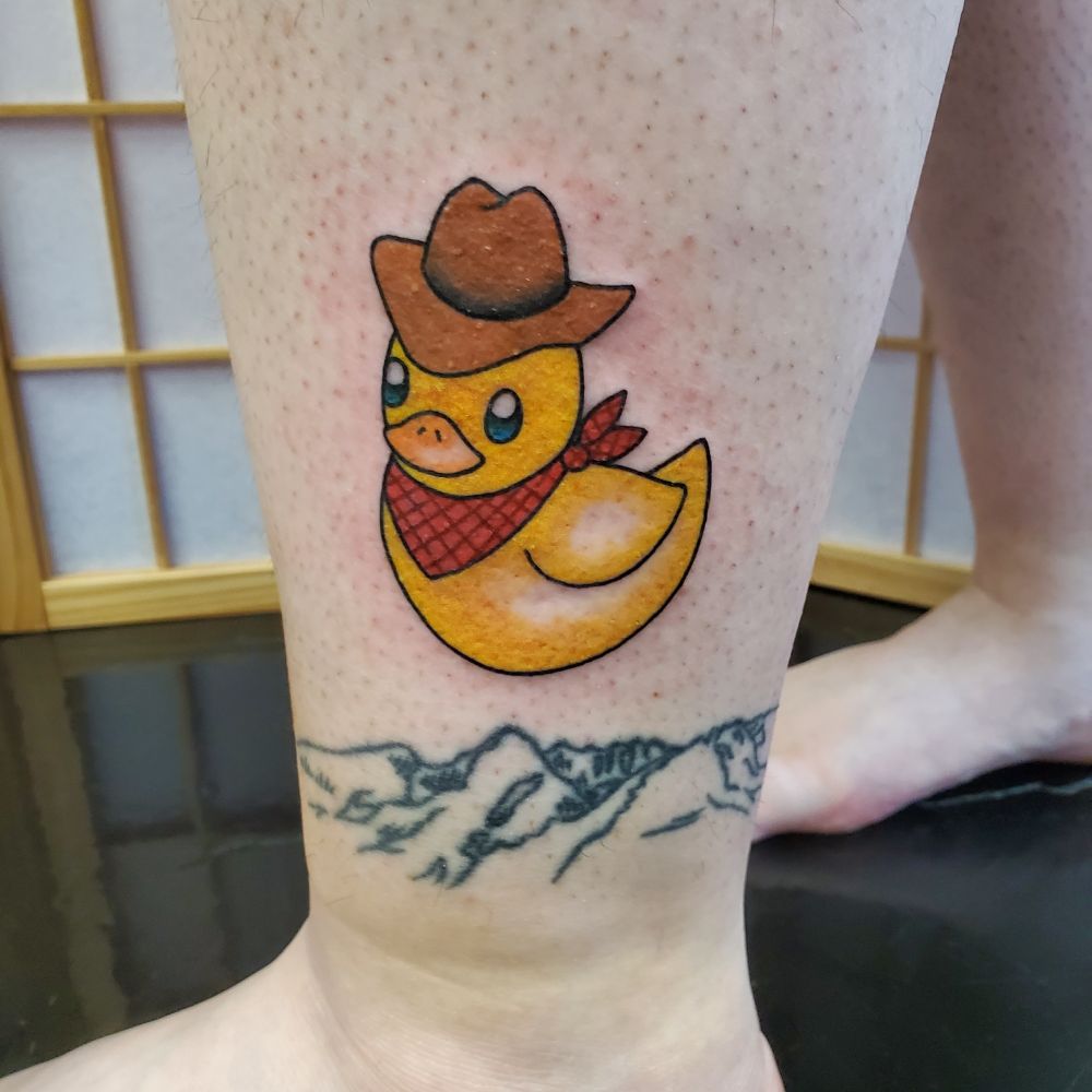 Tattoo on the lower leg of a yellow rubber ducky wearing a cowboy hat and red bandanas 