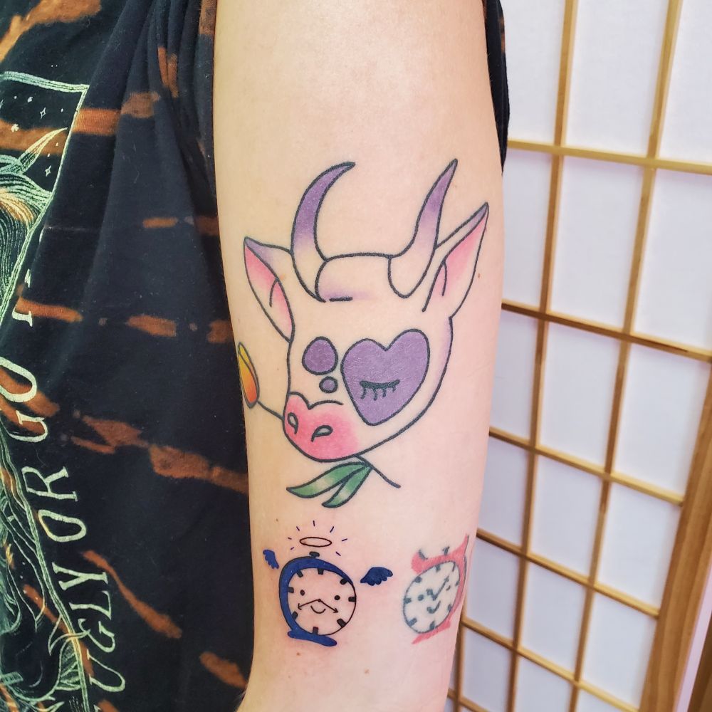 Tattoos on the outer forearm. A larger healed piece of a cows head holding a tulip in their mouth with a heart shaped spot on the face colored in lavender. Beneath are two tattoos of clocks, one blue and with angel wings, one red with devil horns. The red one is an older healed tattoo. The blue is fresh