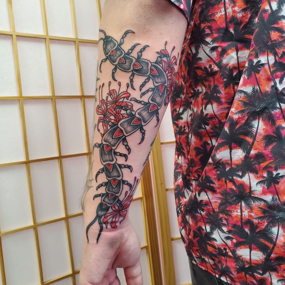 Tattoo of a black centipede on the back of the arm with read spider lily flowers around it