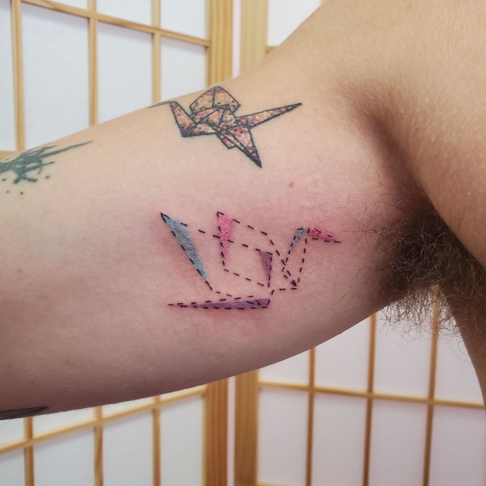Tattoo on the upper inner arm of a dotted line origami crane with a few spots of color in blue pink and purple 