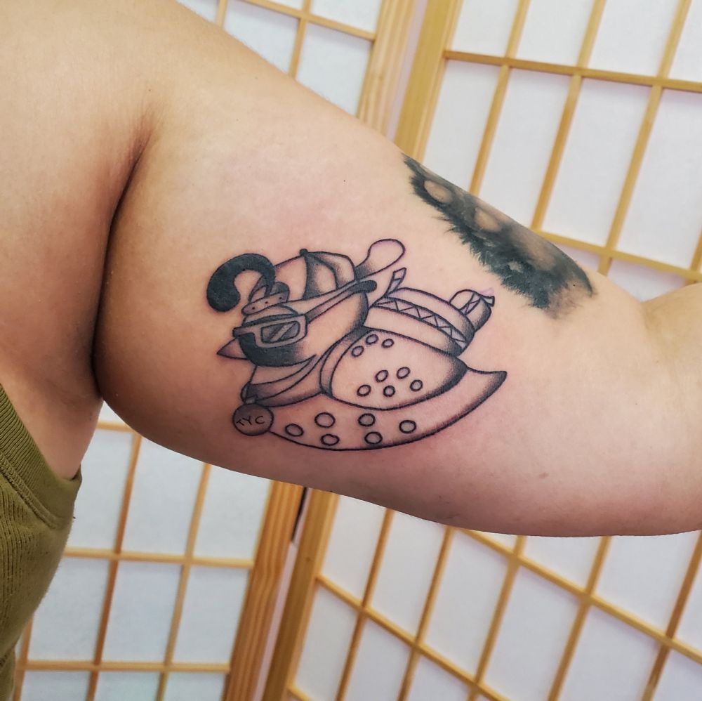Tattoo on the inner upper arm of a quail wearing a backpack, baseball cap and sunglasses, in black and grey