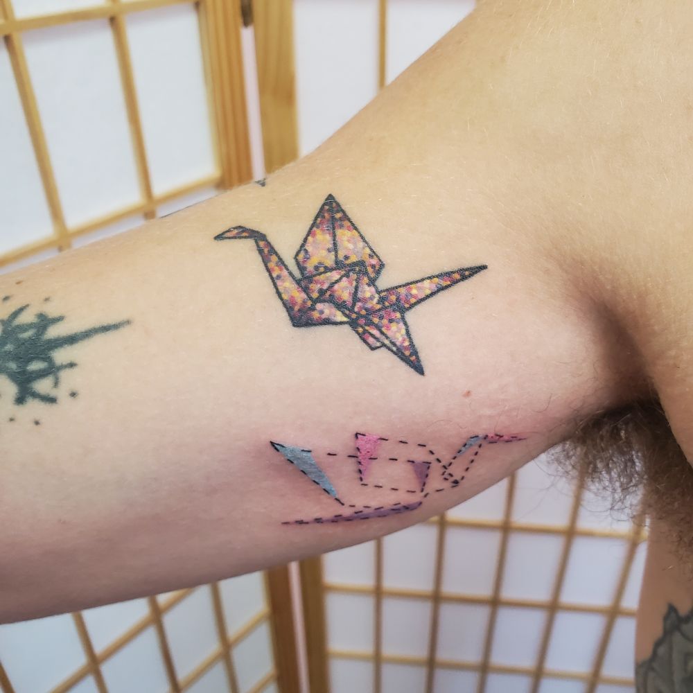 A healed origami crane tattoo on the upper arm. The crane is dotted to look like glitter. It's purple gold and pink 