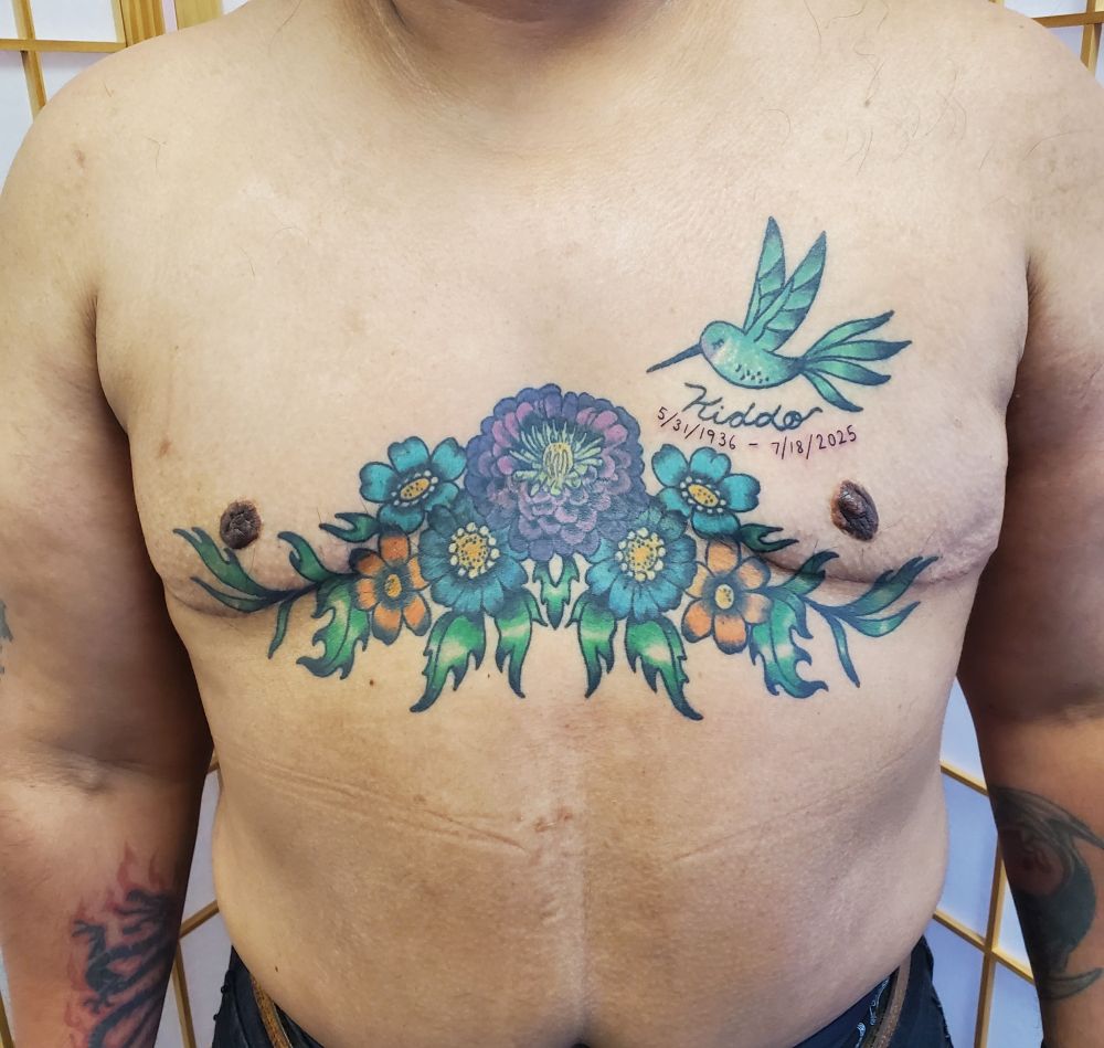 A healed floral chest piece, with a large zinnia flower on the sternum, and daisies and marigolds. A humming bird flies by in the top right. The tattoo is in full color, with blues, purples oranges and greens
