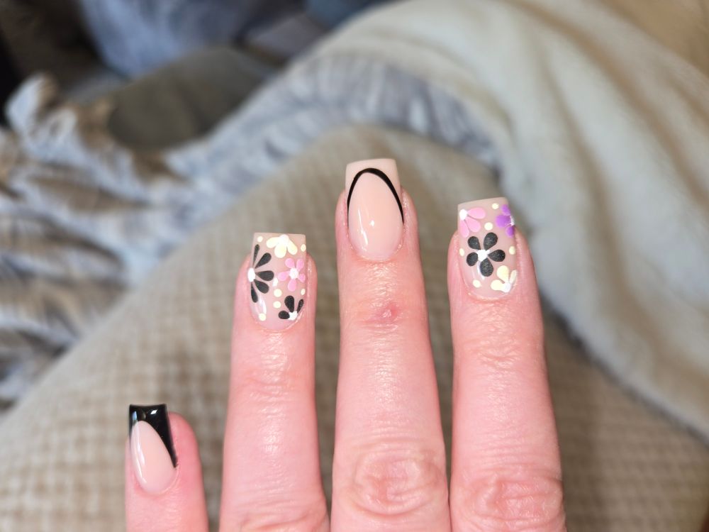 Four fingers. The pinky has a black French tip. The pointer and ring finger have 3d flowers in shades of black, pink, purple, and off white, with some white dots. The middle finger has a black curve that mimics a French tip but the tip of the nail is nude. 