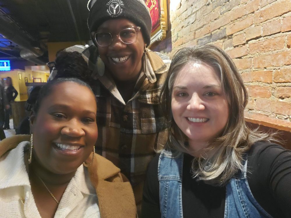 3 people smiling at the camera, 2 with darker skin and 1 with more fair skin.