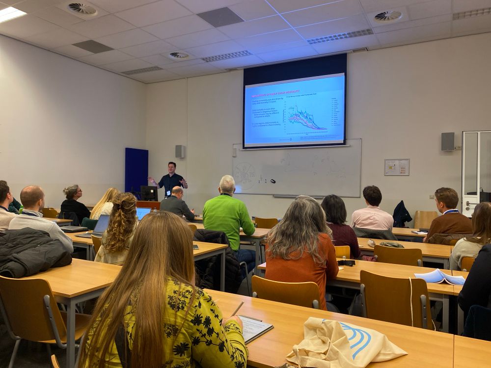 Vibrant male Professor named Tim standing in front of slides projected on to a background wall, there is a crowd in front of him listening intently at an academic conference.  