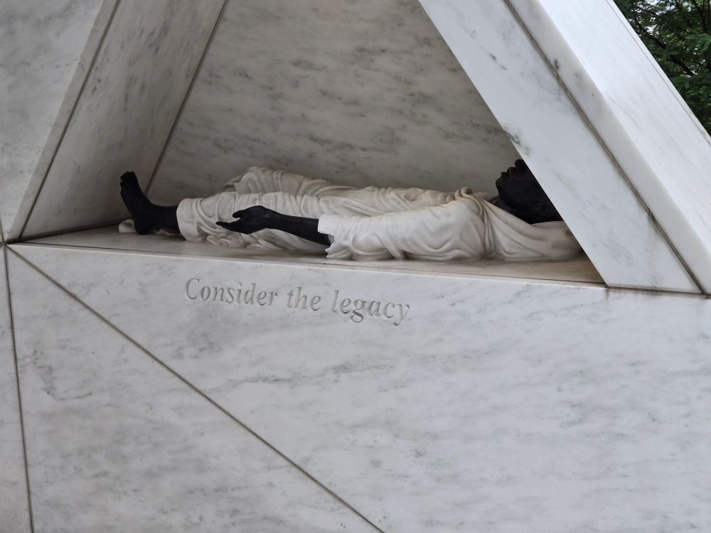 A portion of a marble monument depicting a robed, dark-skinned person lying on their back in a niche, reminiscent of the way enslaved people were chained on transatlantic slave ships, with the words, "Consider the legacy"