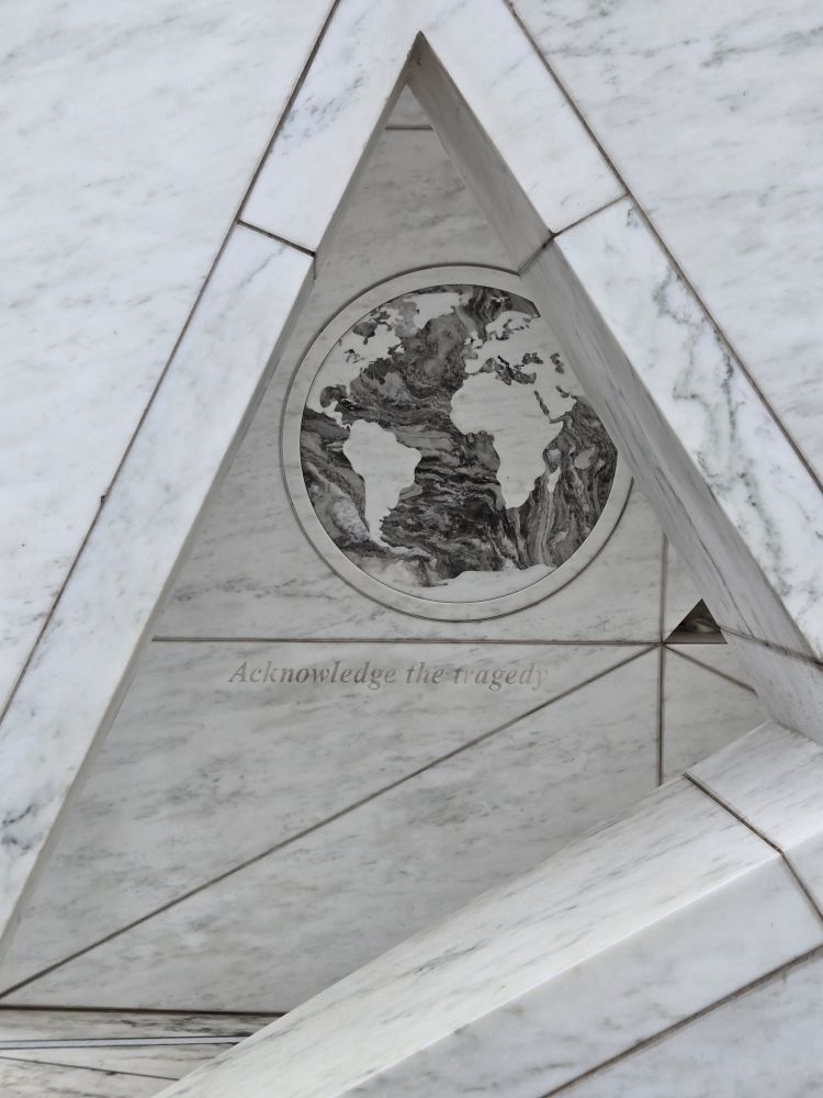 A triangular portion of a marble monument depicting the globe and the words, "Acknowledge the tragedy" 