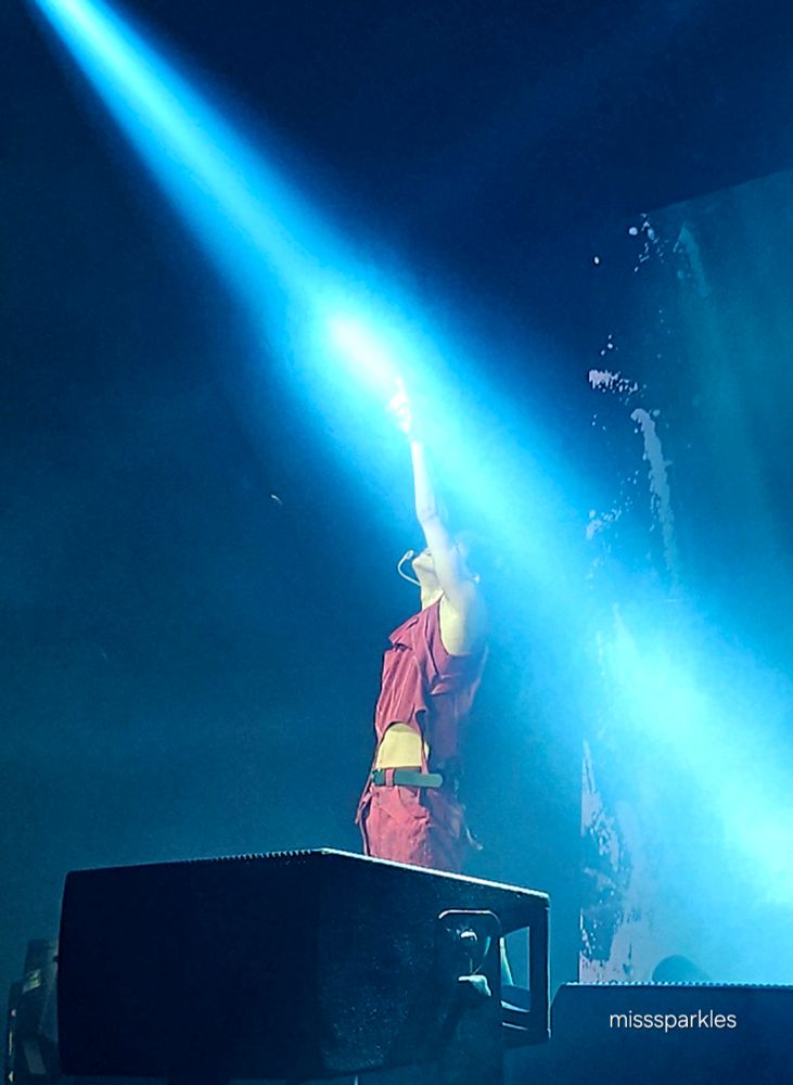 Taemin in the main stage, seen from the side, touching the pearl aqua spot light with his arm outstreched above his head, with gis head tilted up. His red crop top going up and exposing part of his belly
