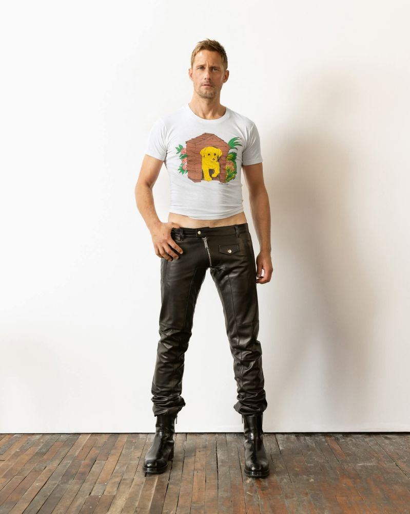 Alexander Skarsgaard modelling for jimmy choo. He is standing in front of a white wall wearing a fitted, cropped, white shirt with a puppy on it, black leather trousers that end at the hips and black leather boots.