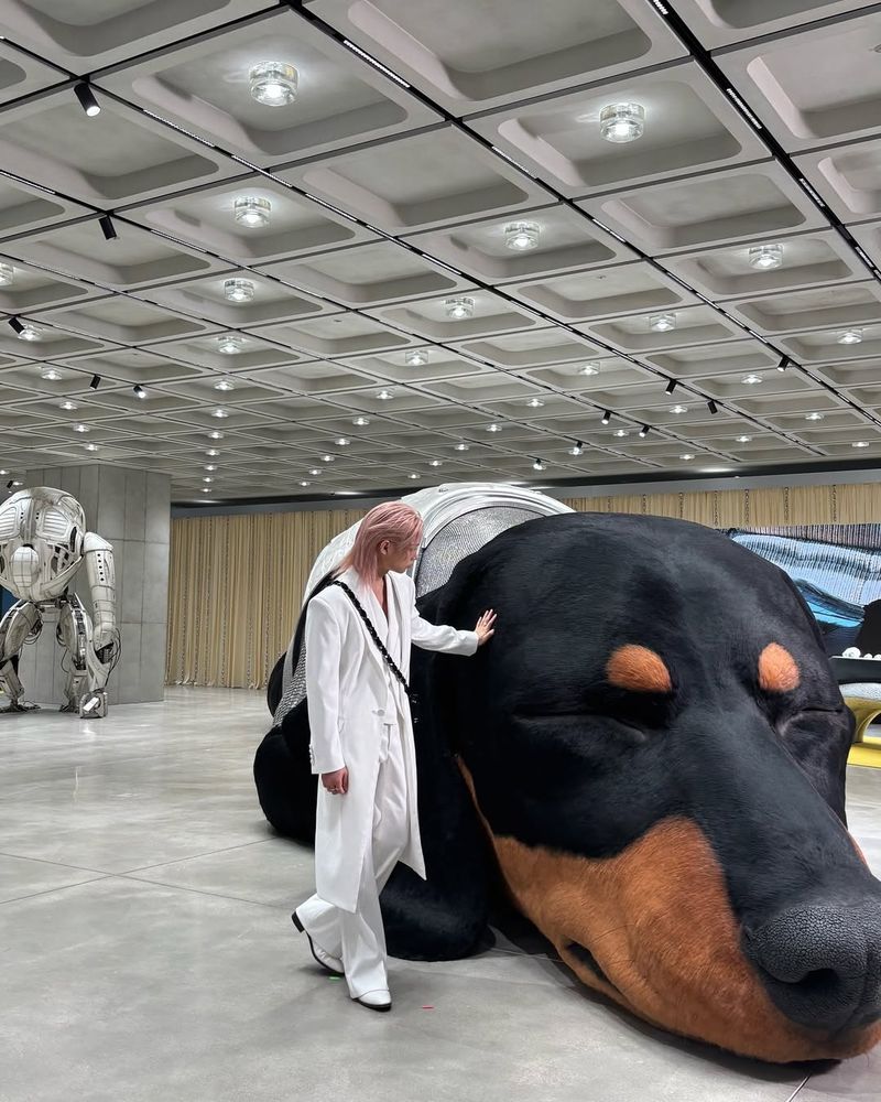 Felix in all white with pink hair standing next of the huge head of a realistic looking dog and gently patting its head