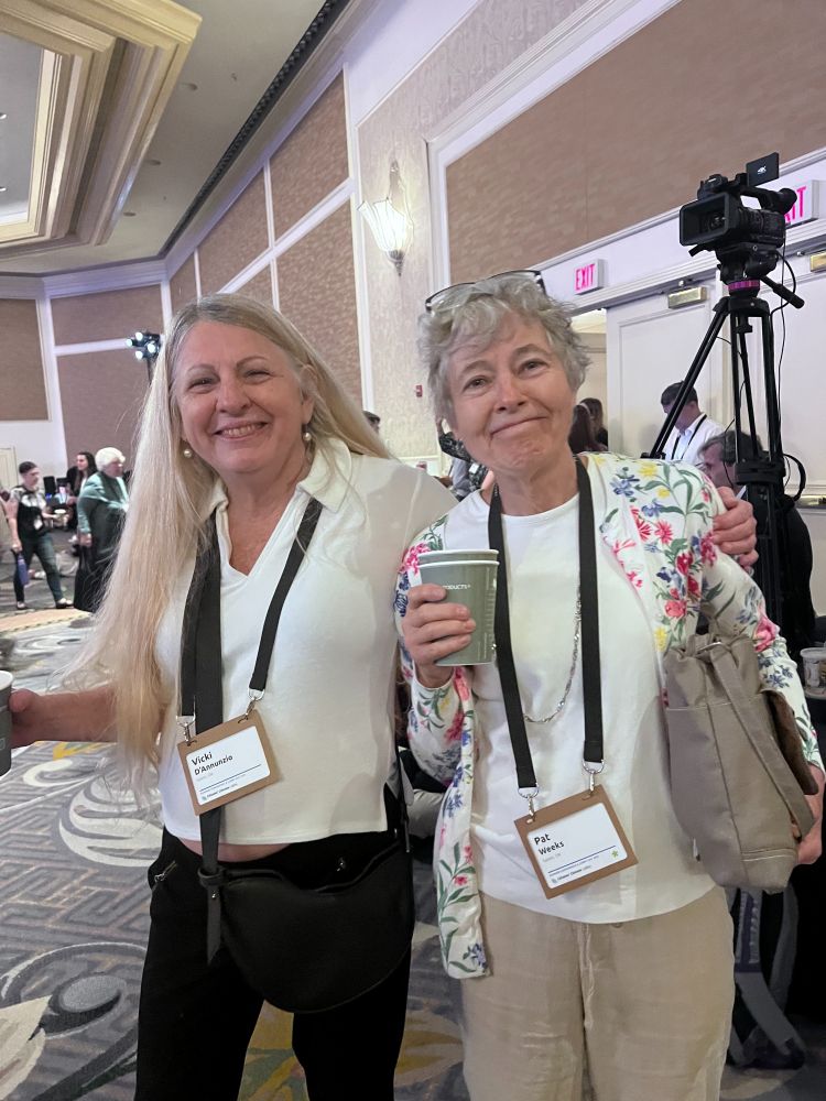 Two older white women wearing lanyards smile at the viewer. They are standing in a hotel ballroom and the background shows other conference attendees walking around.