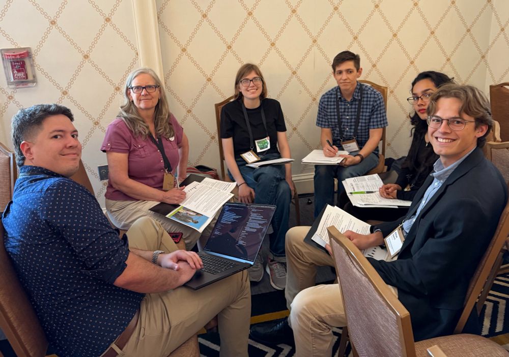A group of six people, different ages and genders, sits in a circle with folders and papers in front of them. They have paused their discussion to smile at the photographer