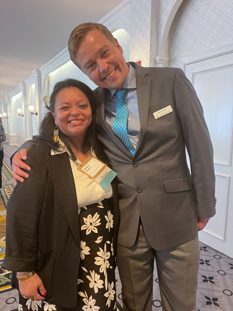 A tall white man in a suit has his arm around a shorter Latina woman in a blouse and floral skirt, and his head is tilted down closer to hers. They both smile at the camera