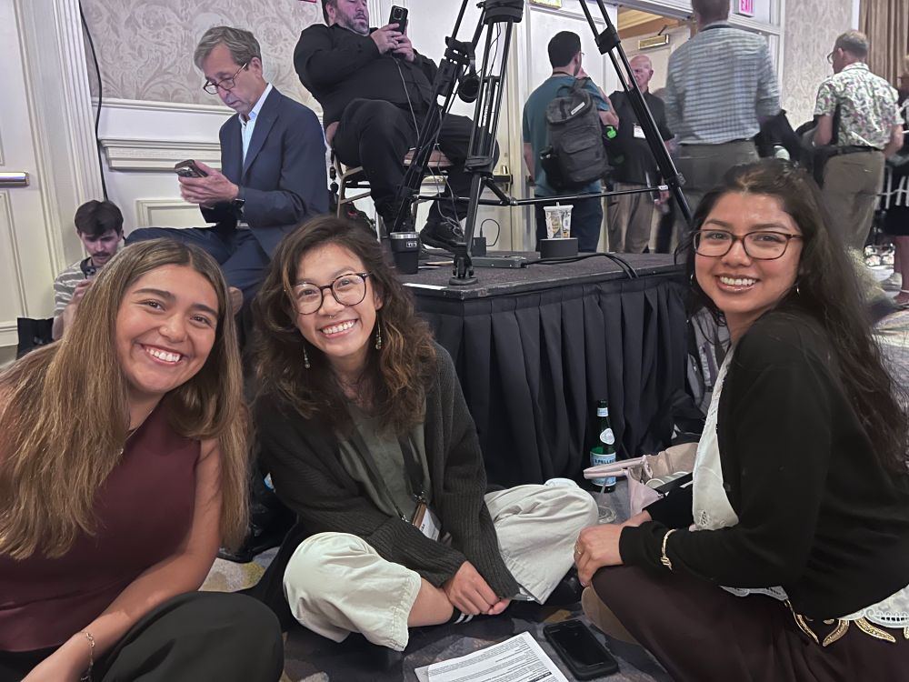 Three young women are seated on the floor of a hotel ballroom in between conference sessions, smiling at the camera, with notes on papers in front of them