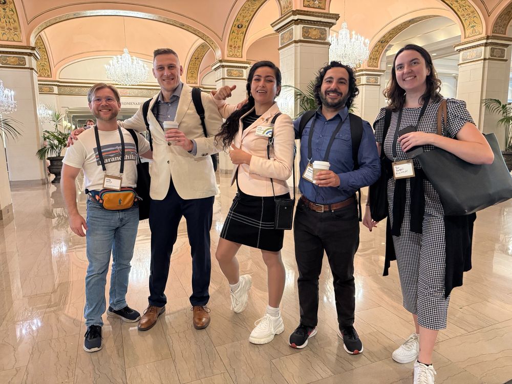 Five young adults wearing conference lanyards stand in a hotel lobby, smiling at the viewer