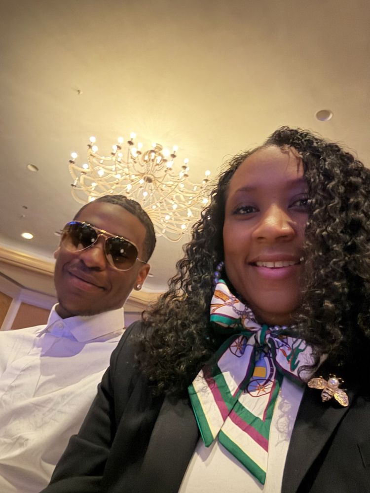 A Black woman, wearing a silk scarf and blazer, and young Black man wearing a button down and sunglasses, are shown in a selfie from the hotel ballroom, smiling at the camera