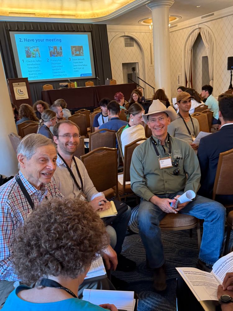 Older white men sit in a semi-circle holding folders and papers. Some are talking to each other, two have looked up and smiled at the photographer. One of the smiling men wears a cowboy hat.