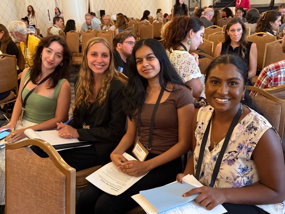 Four young women of different ethnicities are sitting in a row of conference seating. They are wearing nametag lanyards, holding folders with papers on them, and smiling at the viewer