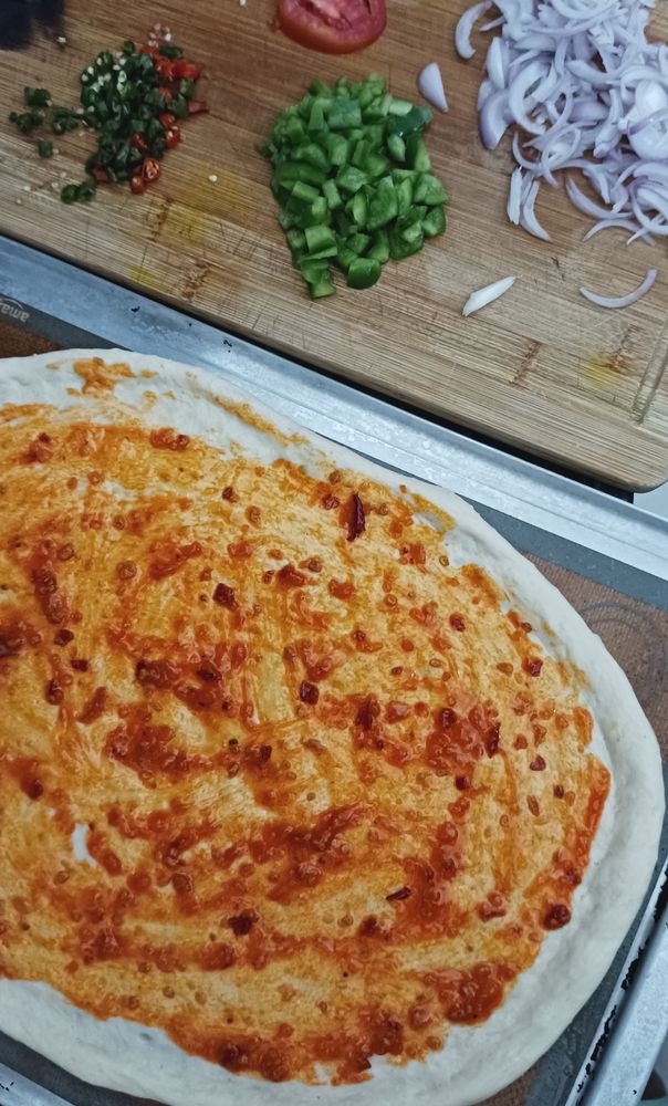 The dough spread out with red sauce applied on it. Above you can see a chopping board with capsicum, chillies, onion and tomato slice. 