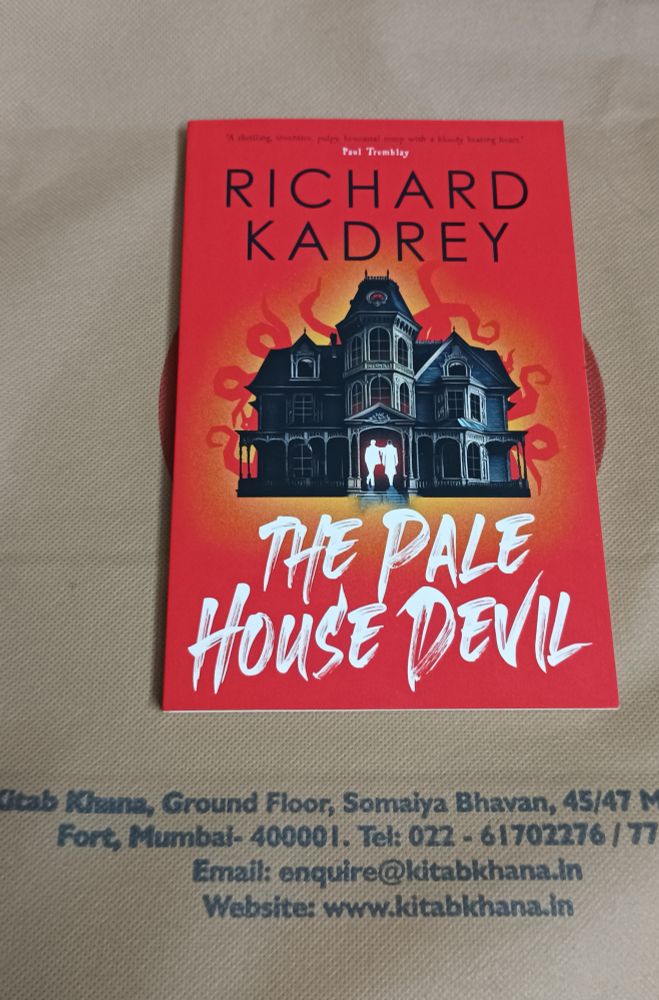 The novella 'The Pale House Devil. It's slom book with a bright red cover. 

On the very top of the cover, there is a quote by author Paul Tremblay written in tiny black letters - "A thrilling, inventive, pulpy, bi-coastal romp with a bloody beating heart. " 

In the middle of the bright red cover is a very dark sinister looking house (mansion?),  behind this house are either red flames or tentacles that seem to frame the house like a foreboding halo. 

Against the dark front door of the house are two white silhouettes, only one of the figures has a black skull...💀


The book is kept on a deep beige carry bag from Kitab Khana bookstore in Mumbai and you can see the address of the store printed on the bag in black. 

"Kitab Khana, Ground Floor, Somaiya Bhavan, 45/47 M. G. Road, Fort, Mumbai- 400001. Tel: 022-61702276/77/78
Email: enquire@kitabkhana.in
Website: www.kitabkhana.in"
