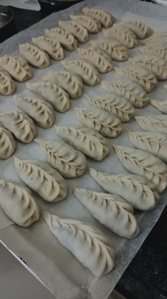 A silver oven tray covered with white parchment paper on which handmade momo (dumplings) are kept in 4 neat rows. 
