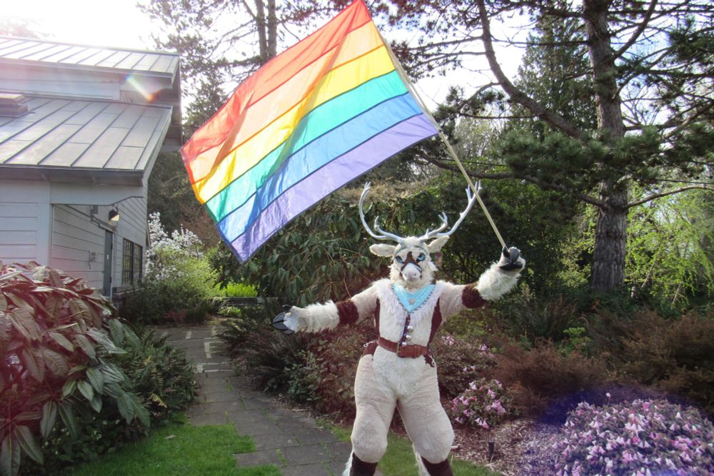 A photograph of a reindeer fursuiter waving a large, rainbow pride flag over their head. 