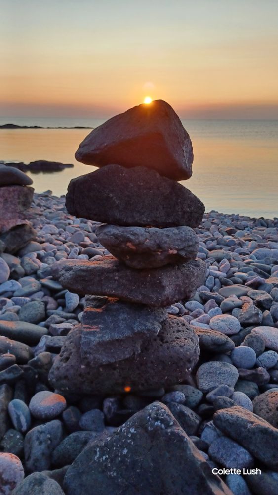 Stacked rocks on a pebble beach with an orange glow from the sun setting behind it. The water in the background is Lake Superior.