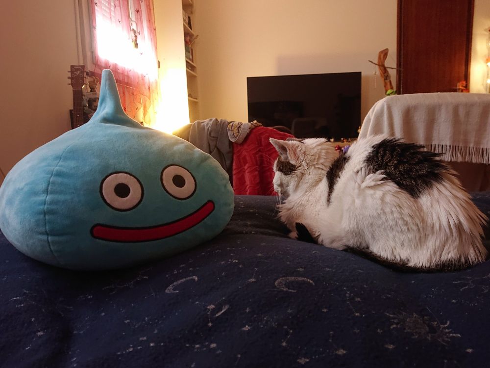 Peluche slime dragon quest plus grande qu'un chat blanc et noir sur un lit. 