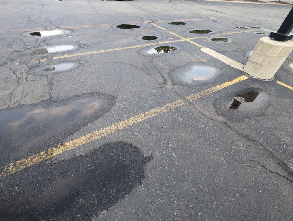 The surface of an empty parking lot showing a few parking spots (outlined in yellow lines), part of a lamp post, and pools of water. The pools of water appear where the wheels of the car would be.