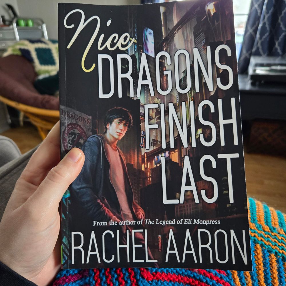 The book cover of Nice Dragons Finish Last by Rachel Aaron. A young white man in casual clothes leans against a wall with a neon-lit cityscape behind him.