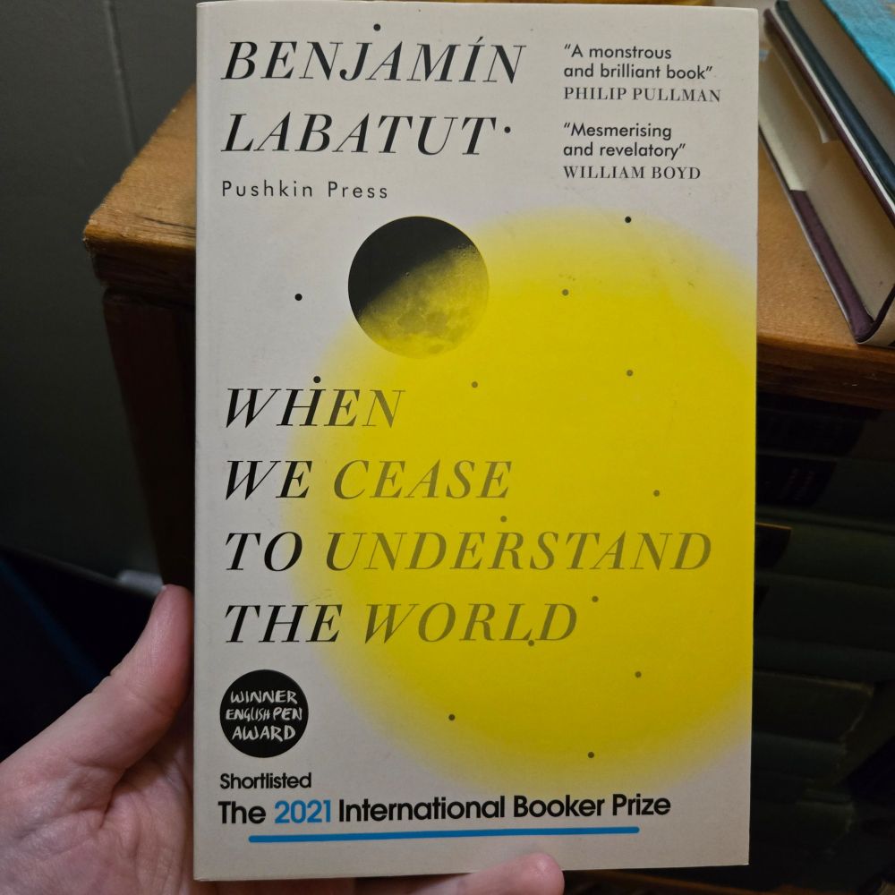 The cover of When We Cease to Understand the World by Benjamin Labatut shows a black circle eclipsed by a semi-translucent yellow circle with fuzzy edges. Where the yellow has covered the black circle, a texture like the surface of the moon shows.