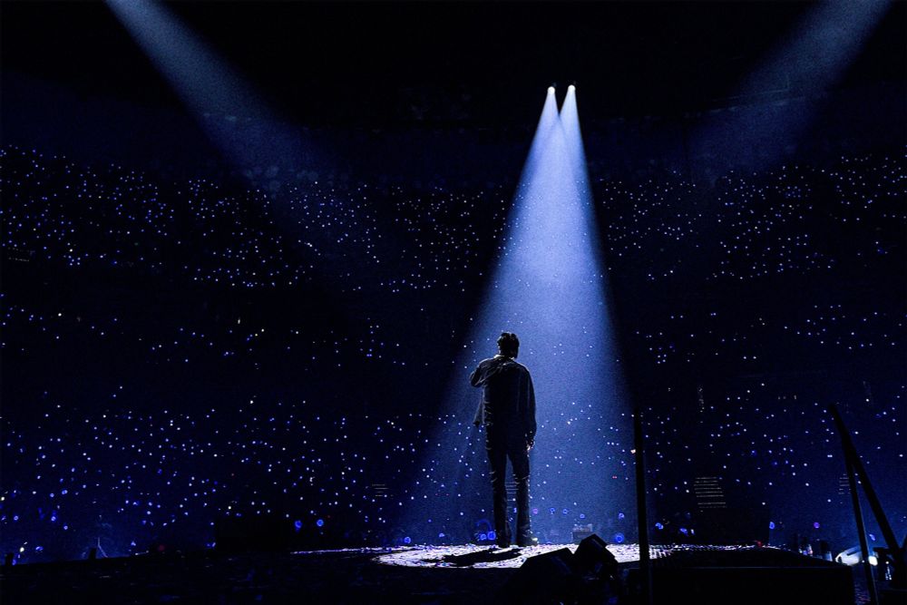 jin of bts standing in the spotlight in one of his concerts with the audience in the dari and lit up lightsticks that make them look like stars.