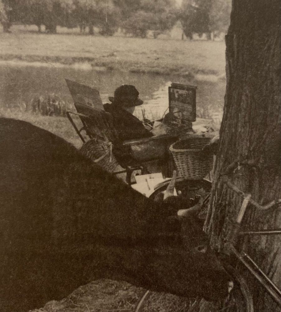 black and white 1954 photograph of a woman painting by the river. She is wearing a black hat and her easel is attached to her wheelchair. In the foreground a cow is investigating a bicycle.