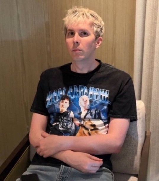 Phil Lester with the dan and phil and a tiny horse shirt on looking...warily?...at the camera