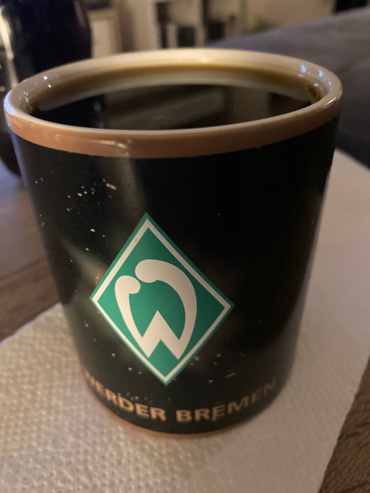 Schwarze Tasse mit grün weißen Werder Logo, randvoll mit schwarzen Kaffee, unter dem Werder Logo steht in Gold Werder Bremen. 
Unter der Tasse ist ein weißes Zewa.
