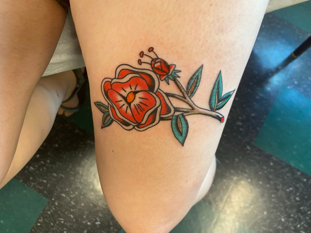 On a woman’s aggressively pale thigh, an American traditional tattoo of a bright red flower on a stem with bluish-green leaves. There’s red at the bottom of the stem, suggesting that the flower bleeds, too. Below her is a black and green linoleum tile floor. 