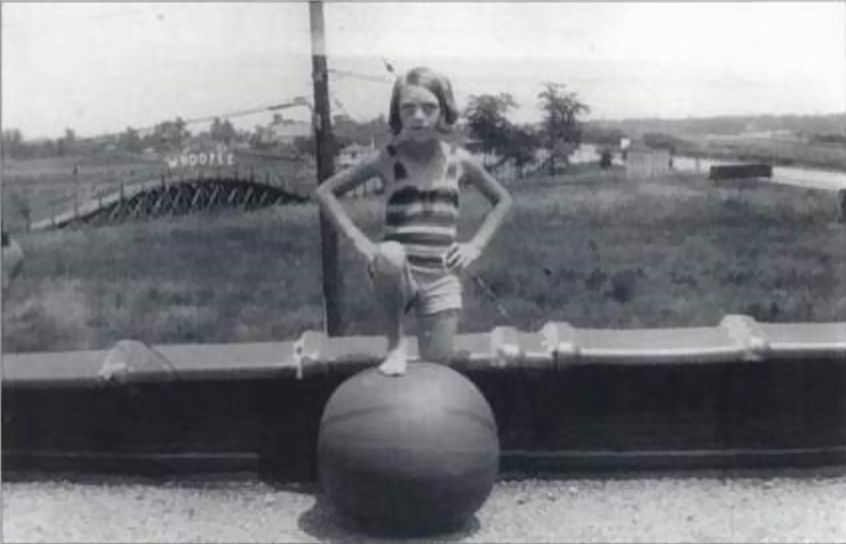 A black and white photo from the 1920s of a young girl standing on a rooftop with her right leg propped up on a large ball. In the background is a whoopie coaster in a field.