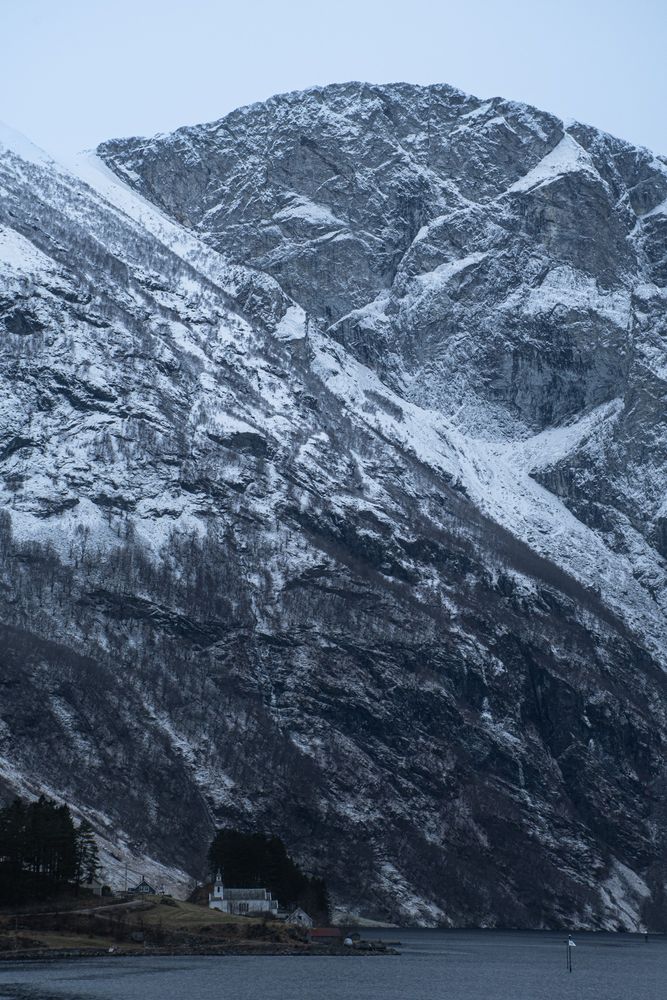 A rocky, snow covered fjord. Light gray, almost white skies. At bottom, very small, is a tiny Norwegian town with a church. 