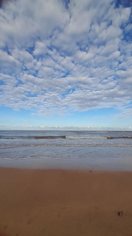 Foto da praia com ondas calmas e uma faixa curta de areia, o céu azul e nuvens brancas e espaçadas 