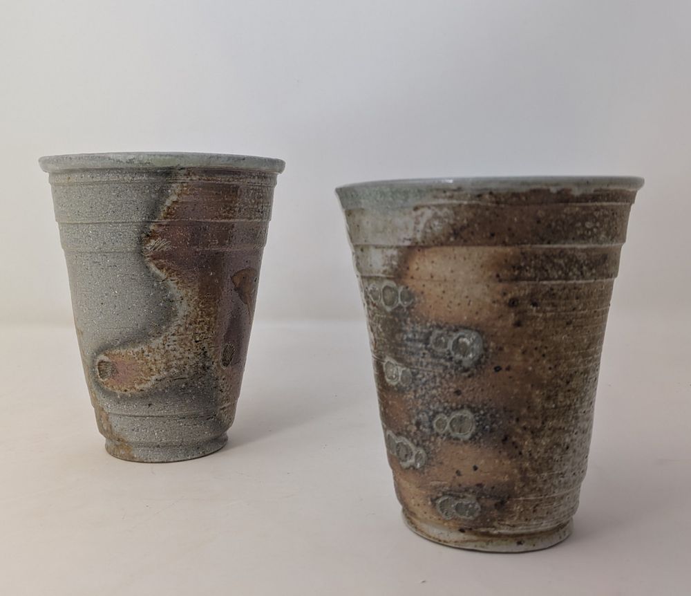 Two ceramic cups both using the shape of a Solo cup. The left wood-fired with markings left by the flame and wood ash. The right soda-fited showing the markings of seashells on the surface.