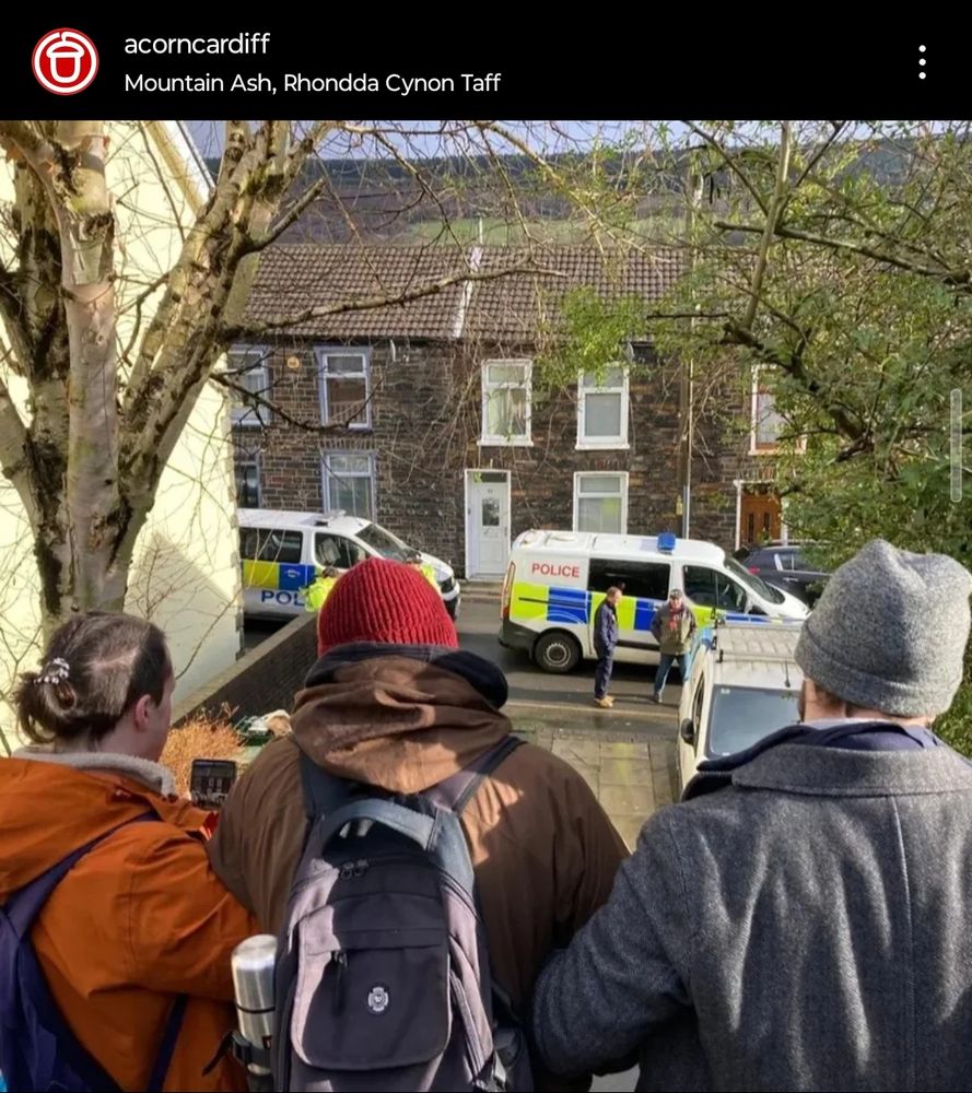 Three ACORN Cardiff members seen from behind arms linked, resisting an eviction, as a significant police presence amasses in the background