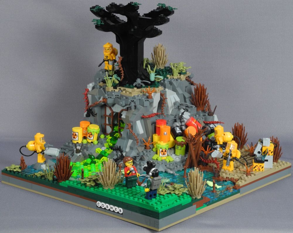 A disturbingly colourful Lego diorama featuring a rocky outcrop from which some kind of industrial effluent discharges into a river. Whatever is flowing out of that pipe is virulently green and most definitely hazardous to any kind of environment. All the fish in the river are dead and most of the plant life has succumbed. There are also a number of chemical drums that have clearly been dumped, bearing radiological and other hazard warnings. Several of the drums are leaking. Fortunately, there is a four person Hazmat team on the case, all properly suited up and toting various detectors and other equipment. From the other side of the river, a reporter is giving a piece to camera. Bricksanto are probably in for some serious fines once news of this gets out.