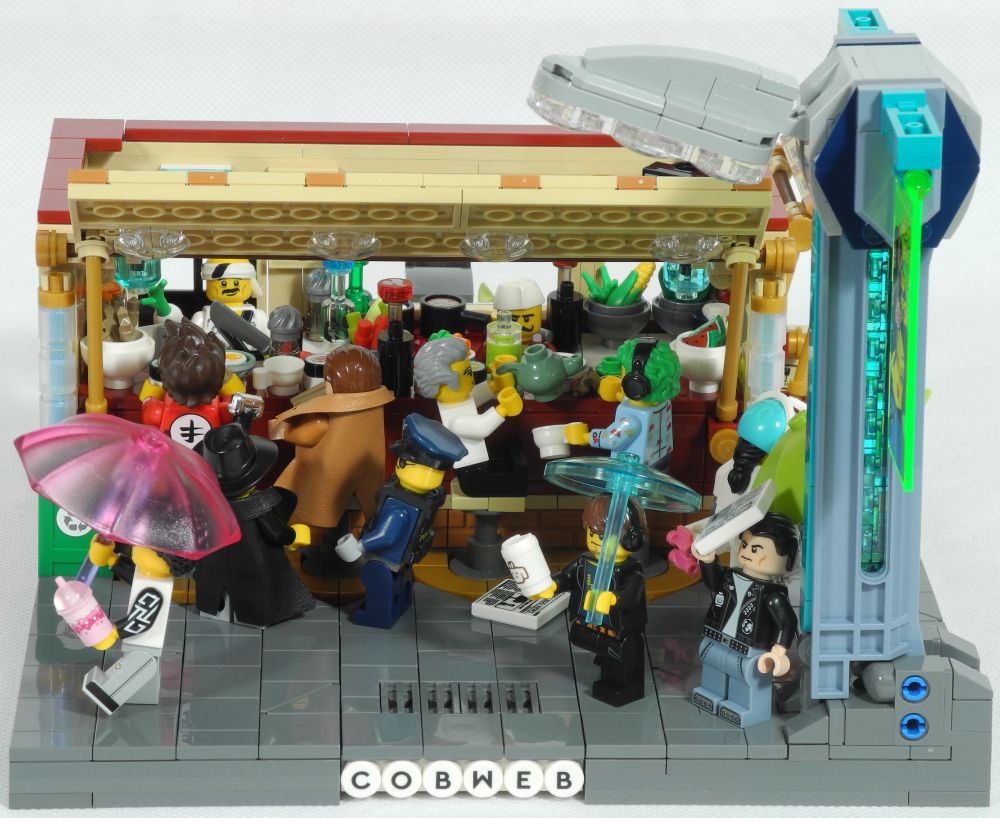 A rather busy little lego diorama of a street-side noodle bar. The two cooks are serving a steady stream of customers, including one who seems to have attracted the attention of a couple of cops, one uniformed, the other in overcoat and fedora. Passers-by have their umbrellas out, apart from one man standing under a street-light, trying to shield himself from the rain with his newspaper.