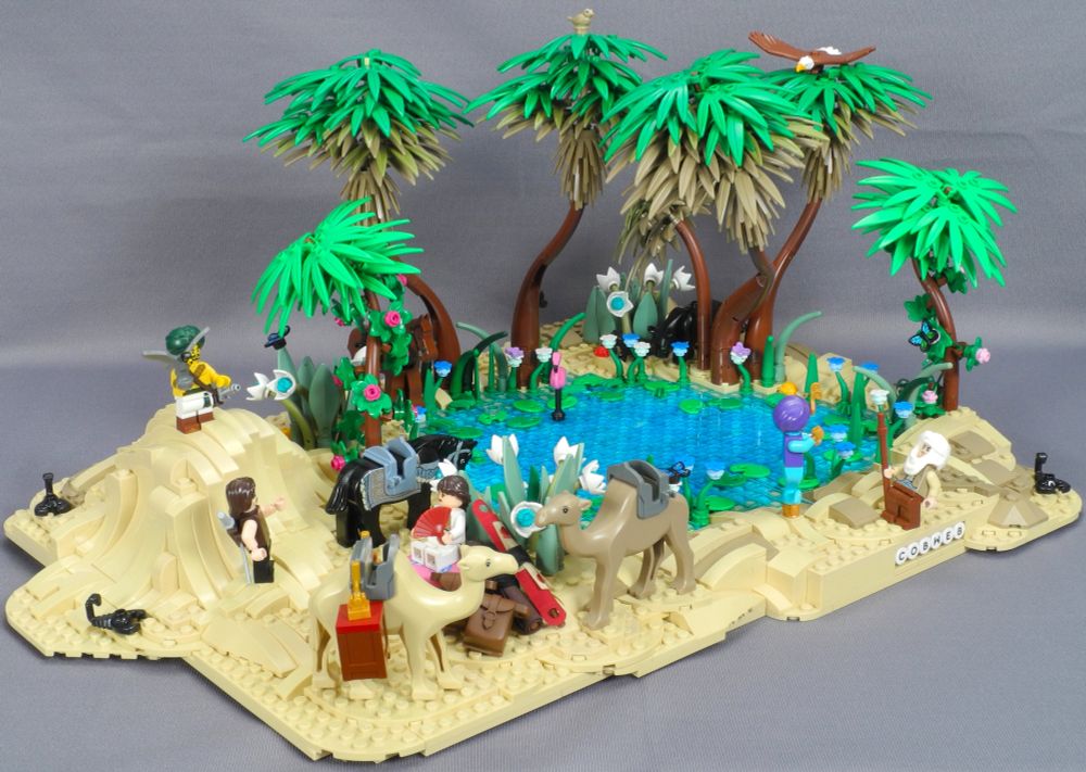 Overview of a lego diorama of a desert oasis. There is a central pool surrounded by lush palm trees and other vegetation. A group of travellers has stopped to rest, their camels unloaded, horses allowed to drink from the clear blue water. A scattering of scorpions, butterflies and other bugs are sprinkled here and there, along with various other bits of wildlife lurking among the plants.
