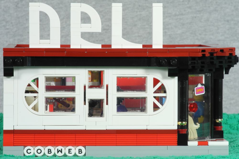 Front view of a small Lego building, in white, red and black, with fancy rounded windows and a corner door with pillars to either side, and a sign above the facing side that says "Deli" in large white free-standing letters.