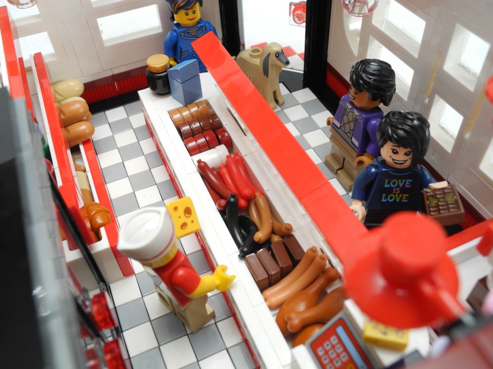 A view across the store, which looks very bright and clean, looking across the central counter towards the door in the far corner. The shielded counter contains various sliced meats, sausages, black pudding and drumsticks which must surely have come from ostriches, given their size relative to the occupants of the store. Beneath a wall-mounted light we can just see the cash register with a note-pad sat on top of it. There are three customers in the store. A lady in a smart blue suit with an Afghan hound, a man (Raj Koothrappali from the Big Bang Theory Ideas set), and an Asian boy, wearing a shirt that says 'Love is Love' in rainbow colours, who looks very pleased to have found some gourmet chocolate.