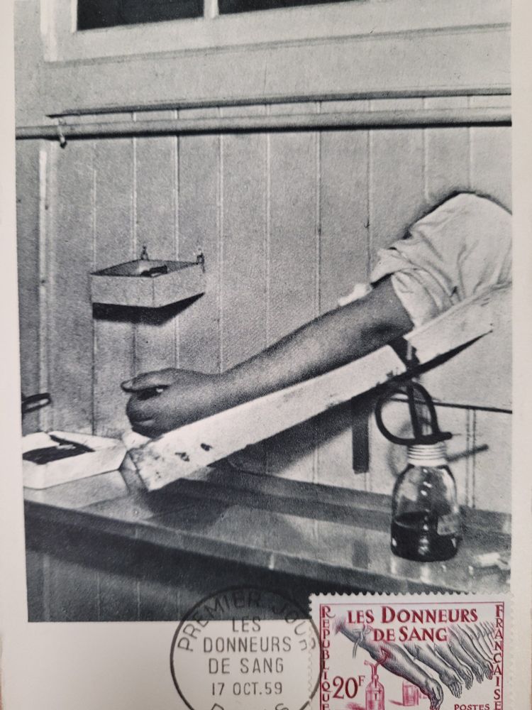 A postcard. A black and white photo shows a disembodied arm sticking through a hole in a wood panelled wall. A length of rubber tube draws blood from the arm into a glass bottle. A red stamp shows a row of identical arms all giving blood into red bottles. The stamp reads "Les Donneurs de Sang" (French for "Blood Donors")