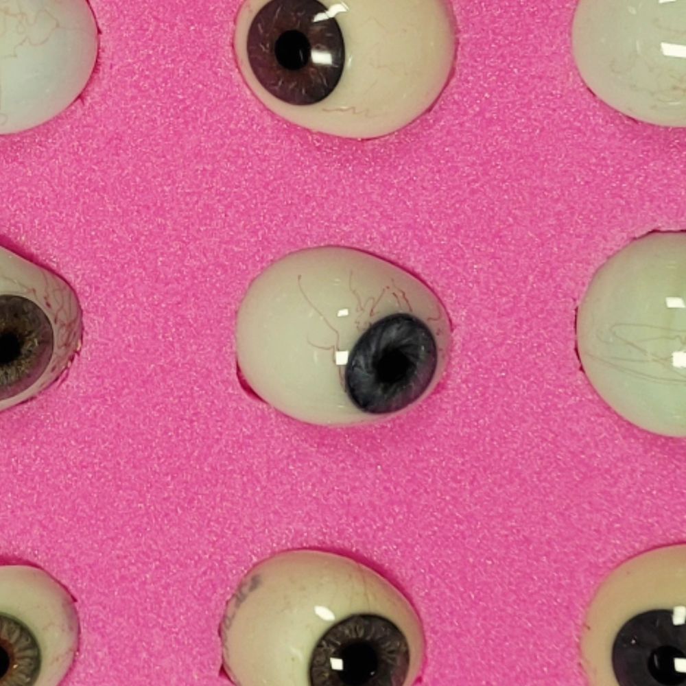 A close up view of a single glass eye with rows of others around the edge of the frame. The eye has a hand-painted dark blue-grey iris and scleral veins made from red thread. It sits on a bed of pink foam.