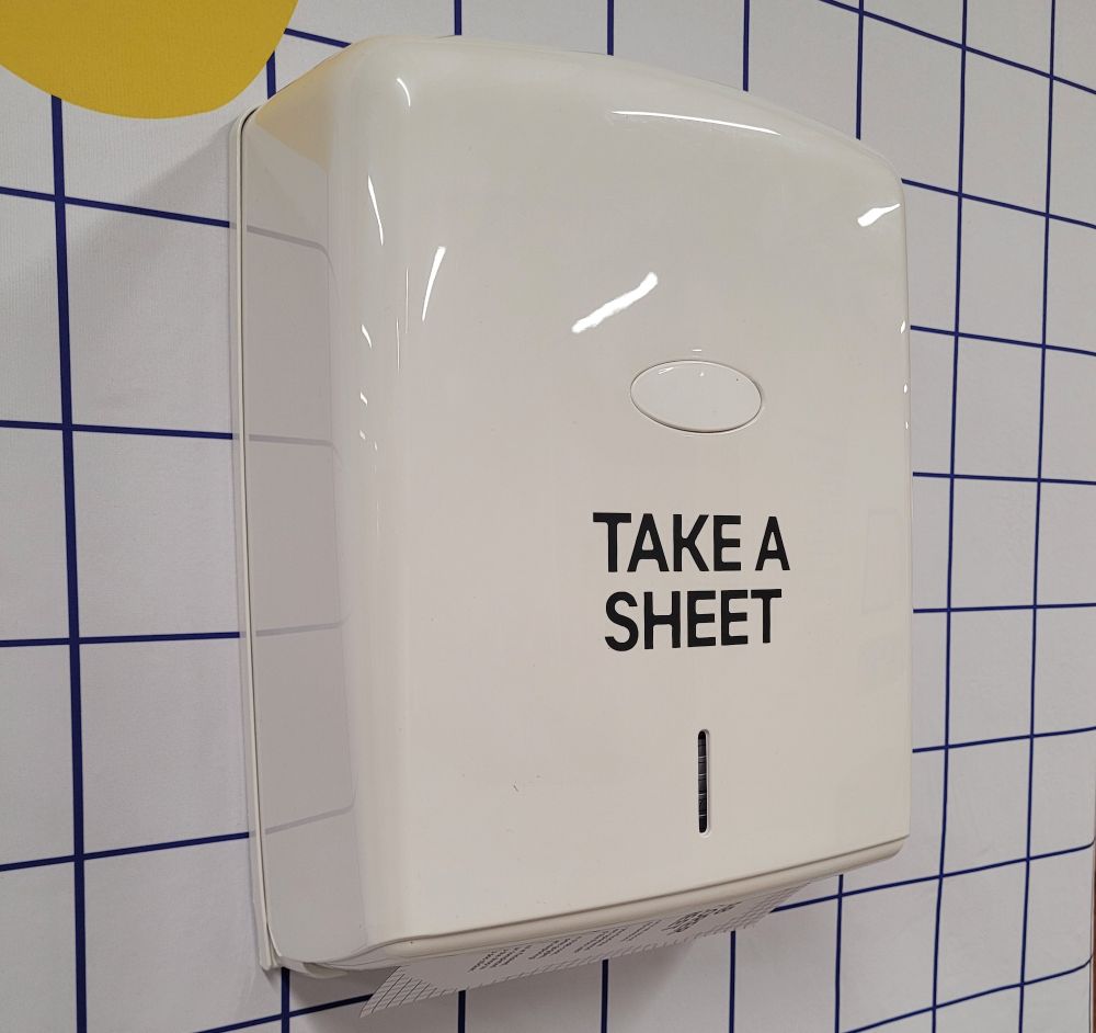 A paper towel dispenser typically seen in public toilets. It is labelled "Take a Sheet" and dispenses fun worksheets for museum visitors